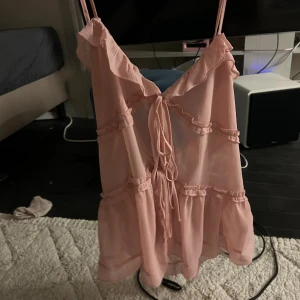 Rosa volangtop med snörning - Supersöt ljusrosa linnetopp med tunna axelband, volangdetaljer och snörning framtill. Toppen är luftig och har en flowy passform med flera volangrader som ger en romantisk vibe. Perfekt för varma dagar och somriga outfits.