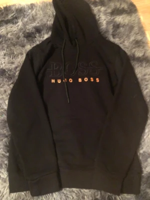 Svart hoodie från Hugo Boss, storlek S - Svart hoodie från Hugo Boss med stor BOSS-logga och orange text på bröstet. Tröjan har huva med snörning och långa ärmar. Mjuk och skön bomullsmix, perfekt för chill dagar. Klassisk passform och stilren design som funkar till allt.