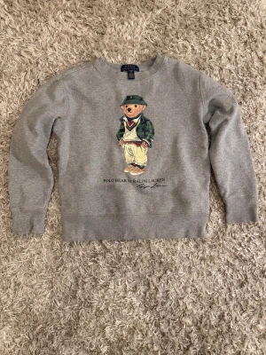 Grå sweatshirt Polo Bear Ralph Lauren - Grå sweatshirt från Ralph Lauren med ikoniskt Polo Bear-tryck på bröstet. Klassisk rund halsringning och långa ärmar. Mjuk bomullskvalitet som är skön att ha på sig. Perfekt för dig som gillar streetstyle och vill ha något med lite attityd. Stl S (8).