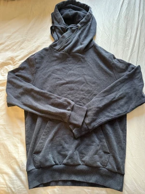 Mörkgrå hoodie med ficka - Snygg mörkgrå hoodie med klassisk känguruficka och ribbade muddar vid ärmar och nederkant. Huva med dragsko för extra stilpoäng. Mjuk och skön bomullsblandning som passar perfekt till jeans eller joggers. Enkel design utan tryck eller logga.