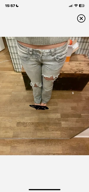 Lågmidjade grå slitna jeans med hål - Säljer ett par grå jeans med slitna detaljer och stora hål vid knäna. Byxorna har låg midja, raka ben och klassiska fickor både fram och bak. Jeansen är i denim och har en cool, avslappnad vibe. Storlek 38 sitter som s
