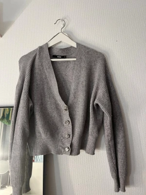 Grå ribbad kofta från Bik Bok - Säljer en grå ribbstickad kofta från Bik Bok i storlek xs. Koftan har v-ringning, långa ärmar och fem knappar framtill. Croppad passform som ger en trendig look. Perfekt att slänga över en t-shirt eller linne för extra lager.