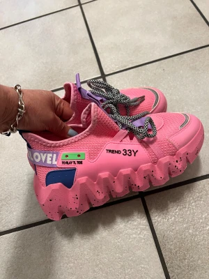 Chunky rosa sneakers med textdetaljer storlek 37 - Säljer ett par riktigt coola chunky sneakers i rosa med lila och blå detaljer. Skorna har svartprickiga snören, grov sula med svarta partier och texten 'LOVE' på hälen. Ovandelen är stickad syntet och har färgglada detaljer som gör dem unika.