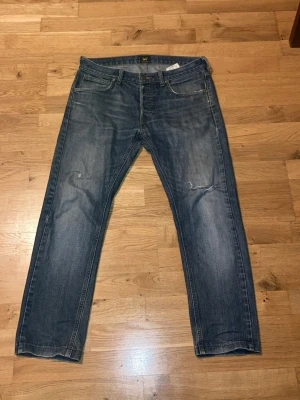 Lee Daren jeans blå W32 L32 - Snygga blå Lee Daren jeans med klassisk femficksdesign och raka ben. Jeansen har en mörkblå tvätt med lätt slitna detaljer och Lee-logga på bakfickan. Passar dig som gillar en tidlös och avslappnad look.