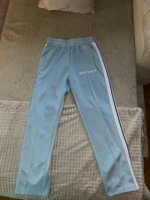 Palm Angels ljusblå mjukisbyxor - Säljer ett par ljusblå mjukisbyxor från Palm Angels med vita sidoränder och logga på vänster lår. Byxorna har elastisk midja med snörning och dragkedjor vid bensluten. Tillverkade i glansig polyester för en sportig vibe.