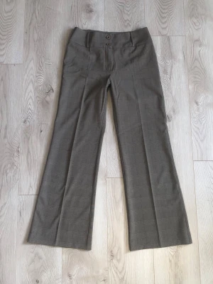 Vintage lågmidjade bootcut kostymbyxor - Snygga bruna kostymbyxor med rutigt mönster, pressveck och bootcut-ben. Byxorna har låg midja och det breda midjebandet med två knappar och hällor ger ett unikt och stiligt uttryck. Materialet är mjukt och bekvämt mot kroppen och är lite stretchingt, vilket gör att de sitter snyggt. Passar storlek 36 och 38. 