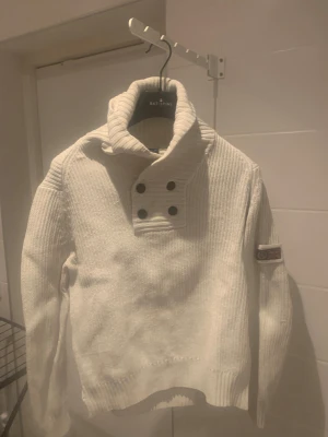 Vit stickad tröja med hög krage Batistini - Vit stickad tröja från Batistini med hög, ribbad krage och fyra svarta knappar framtill. Tröjan har långärmade, ribbade muddar och ett patch med brittisk flagga på ena ärmen. Perfekt för kyliga dagar och har en clean, stilren look.