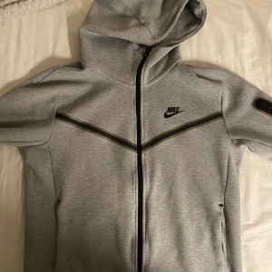 Tracksuit - Snygg grå huvtröja från Nike med dragkedja och huva. Tröjan har svarta detaljer och en klassisk Nike-logga på bröstet. Perfekt för en sportig look eller avslappnade dagar. Tar helst emot byten annars funkar pengar bra också:)