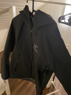 Svart vindjacka från Arc'teryx M - Svart vindjacka från Arc'teryx med huva och dragkedja framtill. Jackan har två sidofickor med dragkedja och justerbar dragsko vid huvan. Perfekt för dig som gillar stilren och funktionell design. Har haft jackan i en vecka säljer den för att det är fel storlek. Priser går att diskutera vid snabb köp 
