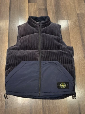 Mörkblå manchesterväst Stone Island - Säljer en mörkblå dunväst från Stone Island i storlek L. Västen har breda manchesterränder, två stora fickor framtill och dragkedja. Klassisk Stone Island-patch på ena fickan och justerbar nederkant. Perfekt för lager-på-lager under höst och vinter.