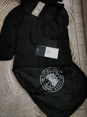 Svart dunjacka från Canada Goose - Säljer en svart dunjacka från Canada Goose med klassisk patch på ärmen och dragkedja framtill. Jackan har en stilren look, huva och flera praktiska fickor. Perfekt för kalla dagar och riktigt snygg till vinteroutfiten.