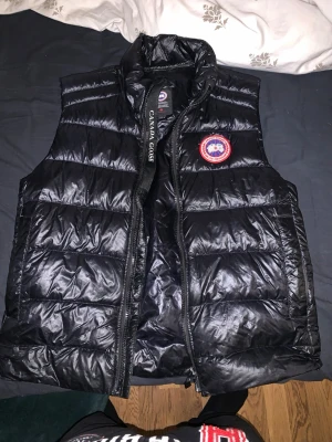 Svart dunväst Canada Goose - Säljer en svart, quiltad dunväst från Canada Goose. Västen har det klassiska runda Canada Goose-märket på bröstet och är tillverkad i glansig polyester. Perfekt för lager-på-lager under kyliga dagar. Västen är i nyskick och köpte den på NK i Göteborg för 6300kr ( pris kan sänkas vid snabba affärer)