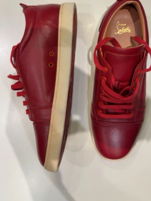 Röda sneakers/skor från Christian Louboutin - Snygga röda sneakers från Christian Louboutin i skinn med matchande röda skosnören. Skorna har en platt sula och klassisk rund tå. Perfekta för dig som vill sticka ut med exklusiva detaljer och stilren design. Du som köpare kan testa få bort märket på sulan men jag har inte testat själv. 