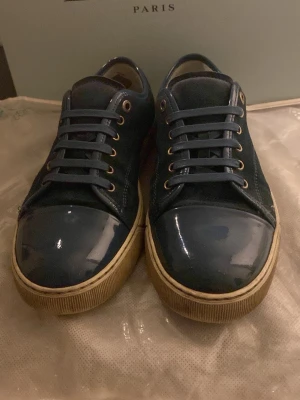 Blå sneakers från Lanvin i mocka och lack (UK6) - Säljer ett par stilrena blå sneakers från Lanvin med snygga lackade tår och detaljer i mocka. Skorna har rund tå, snörning med metallöljetter och en beige sula som ger en cool kontrast. Perfekta för dig som vill ha något unikt och trendigt. Ligger även över 5000kr i ordinarie pris. 