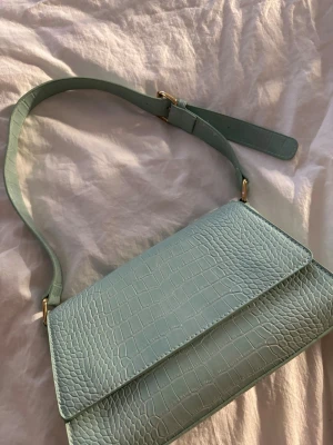 Mintgrön axelväska med krokoprägling - Snygg mintgrön/ ljusblå axelväska med krokopräglad yta och justerbar rem i samma färg. Väskan har guldfärgade metalldetaljer och en stilren, rektangulär form. Perfekt för dig som gillar trendiga accessoarer med lite edge. Knappt använt 