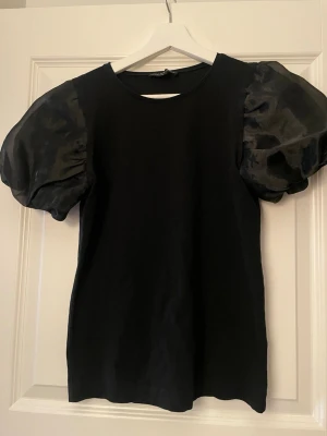 Svart topp med puffärmar - Säljer en svart topp med stora puffärmar i glansigt material. Kroppen är i mjuk bomull och ärmarna ger en riktigt statement-look. Rund halsringning och normal passform. Perfekt för dig som vill ha något extra till outfiten.