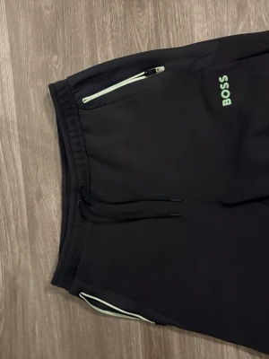 Svarta shorts från BOSS - Svarta mjukisbyxor från BOSS med grön logga på benet och vita dragkedjor vid fickorna. Byxorna har snörning i midjan och är gjorda i mjukt bomullsmaterial. Perfekta för chill dagar eller när du vill ha det bekvämt. Dom är typ helt oanvända då dom var för stor för mig 