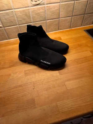 Svarta Speed Trainer svarta sneakers - Säljer ett par svarta svarta  Speed Trainer sneakers i storlek 42. Skorna har en strumpliknande ovandel i stretchig textil, chunky sula och vit -logga på sidan. Riktigt clean och futuristisk look som passar till streetwear.