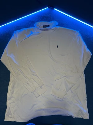 Vit polotröja från Polo Ralph Lauren - Klassisk vit polotröja från Polo Ralph Lauren med broderad marinblå logga på bröstet. Tröjan har lång ärm, ribbade muddar och är gjord i mjuk bomull. Perfekt för lager på lager under höst och vinter. Fläckar finns/ går nog att tvätta 