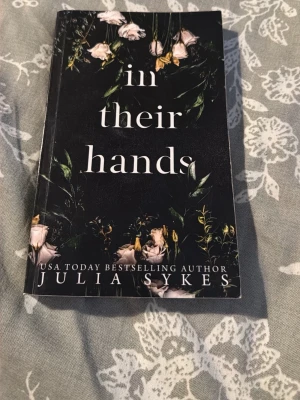 In Their Hands - En spännande bok av USA Today-bästsäljande författaren Julia Sykes. Omslaget har en mystisk känsla med mörka blommor, vilket lockar dig som gillar intensiva och känsloladdade berättelser. Perfekt för unga vuxna som söker något fängslande.