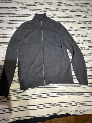 Fullzip merinoull  - Säljer en stilren mörkgrå kofta från Riley i mjuk merinoull. Koftan har hel dragkedja framtill, hög krage och långa ärmar. Perfekt för lager på lager under kyliga dagar och riktigt skön att ha på sig.