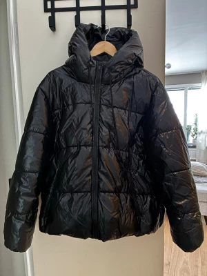 Svart pufferjacka från Vila - Svart pufferjacka från Vila i storlek 38. Jackan har en stor huva, dragkedja framtill och snörning i nederkant för extra komfort. Materialet är glansigt och syntetiskt, perfekt för kalla dagar. Enkel och clean design som funkar till allt.