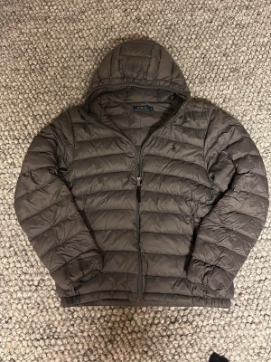 Mörkgrå pufferjacka från Polo Ralph Lauren - Säljer en mörkgrå pufferjacka från Polo Ralph Lauren i storlek L. Jackan har huva, dragkedja framtill och klassisk quiltad design. Perfekt för kyliga dagar och riktigt skön att bära. Enkel och stilren look med diskret logga på bröstet.