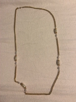 Guldigt halsband med pärldetaljer - Säljer ett långt halsband med grov guldfärgad kedja och vita pärlliknande detaljer. Kedjan har fyra större oregelbundna pärlor och mindre runda pärlor mellan länkarna. Perfekt accessoar för att lyfta din outfit.