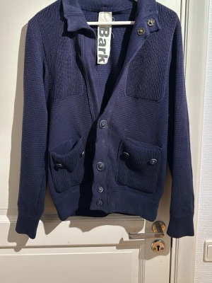 Unik mörkblå cardigan - Säljer en mörkblå kofta från Bark med grovstickad struktur, stora knappar framtill och två fickor med knappdetaljer. Koftan har krage och lång ärm, perfekt för lager på lager under kyliga dagar. Klassisk och stilren design som funkar till mycket.