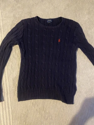 Mörkblå kabelstickad tröja Polo Ralph Lauren - Kabelstickad mörkblå tröja från Polo Ralph Lauren med klassisk röd logga broderad på bröstet. Tröjan har rund halsringning, ribbade muddar och är långärmad. Perfekt för dig som gillar preppy stil.