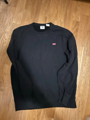 Svart sweatshirt från Levi's - Klassisk svart sweatshirt från Levi's med rund hals och diskret röd logga på bröstet. Tröjan har långa ärmar och ribbade muddar vid ärmslut och nederkant. Perfekt till chill dagar och enkel att matcha med allt.