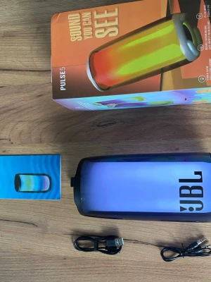 JBL Pulse 5 - JBL Pulse 5 bärbar Bluetooth-högtalare med imponerande 360° LED-ljuseffekter och kraftfullt ljud. Upp till 12 timmars batteritid, vattentät design och trådlös anslutning. Komplett med originalkartong, laddkabel och ljudkabel. Mycket fint skick, inga synliga defekter.