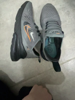 Nike Air 270 grå sneakers - Säljer ett par grå Nike Air 270 sneakers med svart och orange detalj på sidan och logga på plösen. Skorna har snörning, ventilerande meshmaterial och en hög, svart sula med synlig Air-enhet. Perfekta för dig som gillar sportig och modern stil.