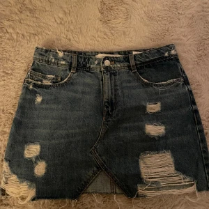 Blå jeanskjol från Zara  - Säljer en blå jeanskjol från Zara  med slitna detaljer och rå kant nertill. Kjolen har klassisk femficksmodell, knapp och dragkedja framtill samt en cool asymmetrisk skärning. Perfekt för dig som gillar en edgy och avslappnad stil.