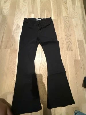 Svarta bootcut byxor från Lindex - Säljer ett par svarta bootcut byxor från Lindex i storlek 134. Byxorna har klassisk femficksdesign, bälteshällor och knapp framtill. Materialet är mjukt och stretchigt, vilket gör dem riktigt sköna att bära. Perfekta för  dig som gillar en tidlös look med lite utsvängda ben.