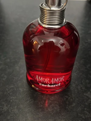 Amor Amor Cacharel 100ml - Upptäck Amor Amor från Cacharel – en ikonisk parfym i röd flaska med modern design. Perfekt för dig som vill sticka ut och addera en touch av romantik till din stil. Flaskans design har en vit ros på baksidan. Volym och ingredienser syns ej tydligt på bilderna.