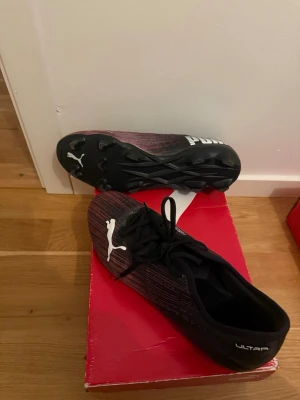 Puma Ultra fotbollsskor svart/röd - Säljer ett par Puma Ultra fotbollsskor i svart och mörkröd med vita detaljer och logga. Skorna har snörning och en lätt, syntetisk ovandel samt dobbar för bra grepp på planen. Perfekta för dig som vill ha snabba och lätta skor till matchen.