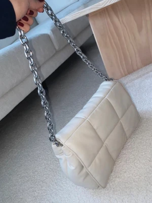 Beige quiltad axelväska med kedja - Säljer en beige quiltad axelväska från HM med chunky silverfärgad kedja som axelband. Väskan har en mjuk, vadderad look och rektangulär form. Använd max 4-5 gr. 
