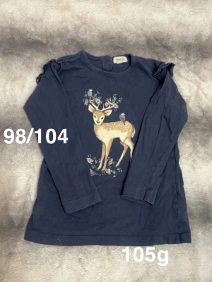 Mörkblå långärmad t-shirt med rådjur - Supersöt mörkblå långärmad t-shirt från Newbie med ett gulligt rådjur och blommor framtill. Små volangdetaljer på axlarna och knäppning bak i nacken. Perfekt för dig som gillar djurmotiv och mjuka material.🌸 Fint använt skick. Storlek 98/104