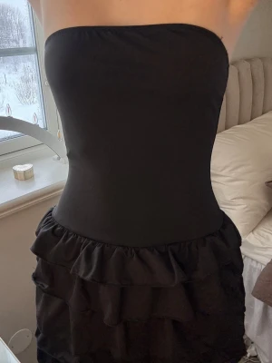 Svart axelbandslös volangklänning - Säljer en svart kort axelbandslös klänning med flera lager volanger på kjolen. Klänningen har en tight passform upptill och är perfekt för dig som gillar en enkel men ändå statement look. Materialet känns mjukt och följsamt.