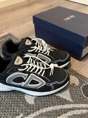Svarta Dior sneakers med vita detaljer - Snygga svarta Dior sneakers med vita och grå detaljer samt stora CD-loggor på sidorna. Skorna har vit snörning, rund tå och en chunky vit och svart sula. Ovandelen är i mesh och syntetmaterial för en sportig vibe. Perfekta för dig som vill sticka ut med lyxig streetstyle.