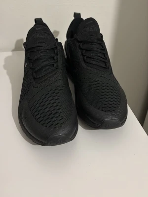 Svarta Nike Air Max 270 sneakers - Säljer ett par helsvarta Nike Air Max 270 sneakers med mesh-ovandel och stor synlig Air-enhet i hälen. Skorna har snörning, diskret Nike-logga på sidan och en sportig vibe. Perfekta för dig som gillar stilrena och bekväma sneakers.
