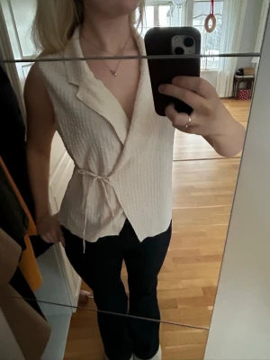 Beige omlotttopp med knyt - Säljer en beige väst med omlottmodell och knyt i sidan. Västen har en lätt strukturerad yta och bred krage, vilket ger en stilren och modern look. Perfekt att styla över en enkel topp eller t-shirt.