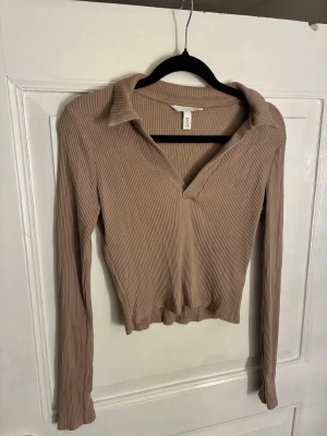 Beige ribbad långärmad topp H&M - Säljer en beige ribbad topp från H&M med lång ärm och krage. Toppen har v-ringning och är croppad i modellen, vilket ger en snygg och avslappnad look. Perfekt att styla med jeans eller kjol för en enkel men trendig outfit. Storlek S men passar XS/S.