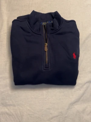 Marinblå half zip från Polo Ralph Lauren - Snygg marinblå tröja från Polo Ralph Lauren med klassisk röd logga på bröstet. Half zip-dragkedja med läderdetalj och hög krage. Tillverkad i mjuk bomull och har långa ärmar med ribbade muddar. Perfekt för en clean och stilren look.