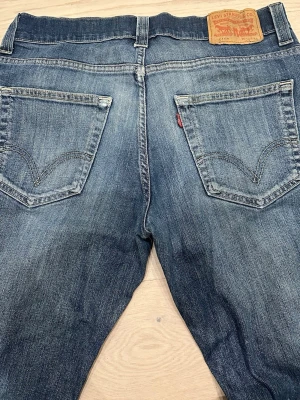 Vintage Levi's 511 blå jeans W36 L34 - Säljer ett par klassiska Levi's 511 jeans i blå tvätt med snygg slitning. Modellen har normal passform och raka ben, fem fickor och dragkedjegylf. Ikonisk läderpatch bak i midjan och röd Levi's-taggen på bakfickan. Perfekta till sneakers och hoodie.