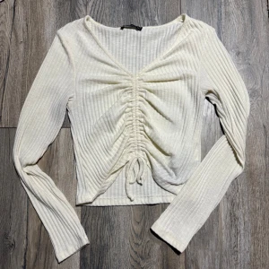 Ribbad vit topp med snörning - Säljer en ribbad vit långärmad topp i storlek M. Toppen har v-ringning och snygg snörning framtill som ger en rynkad effekt. Materialet är mjukt och stretchigt, perfekt till jeans eller kjol. Enkel men trendig design som passar till mycket.