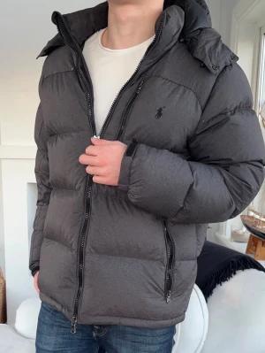 Mörkgrå pufferjacka från Polo Ralph Lauren - Säljer en mörkgrå pufferjacka från Polo Ralph Lauren med huva och dragkedja framtill. Jackan har två sidofickor med dragkedja och diskret logga på bröstet. Perfekt för kalla dagar och riktigt skön att bära över en hoodie eller tröja.