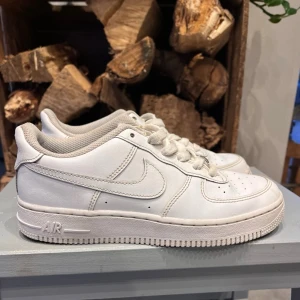 Nike Air Force 1 vita sneakers - Klassiska Nike Air Force 1 sneakers. Skorna har snörning, diskret Nike-logga på sidan och AIR-detalj på hälen. Perfekta för dig som gillar tidlös streetstyle och vill ha en ikonisk sneaker i garderoben.