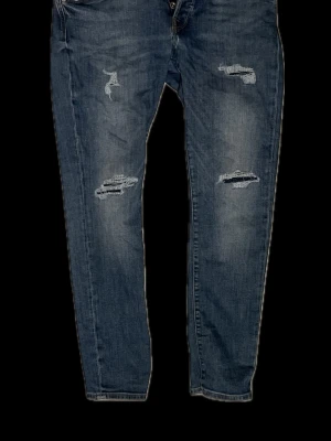 Blå slitna jeans slim fit, strl 30 - Säljer ett par blå jeans med snygga slitningar och raka ben. Jeansen har klassisk femficksdesign, knappgylf och slitna detaljer framtill för en avslappnad look. Perfekta för dig som gillar en casual och trendig stil. Väldigt ovanliga att hitta i butik 